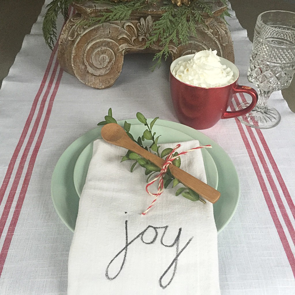 Green plate with folded napkin that says joy along with a cup of cocoa and whip cream