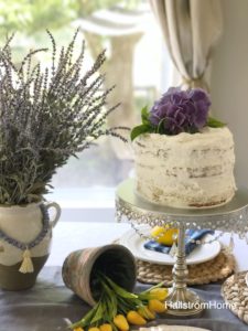 Frosted cake with fresh flowers on top and a cake stand with a vase of lavender next to it