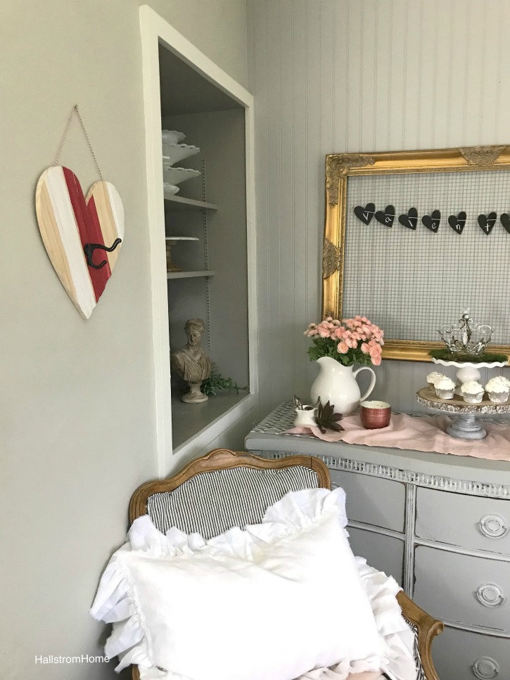 heart shaped coat hook with chair and white linen pillow. gold chicken wire frame on wall with valentines heartbanner and vase of pink flowers. 