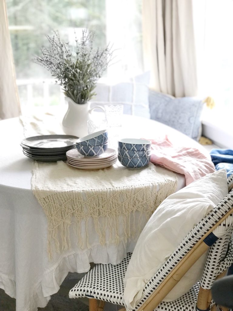 white linen ruffle table cloth with cream tassel table runner. white pitcher filled with lavendar. stack of gray plates and stack of pink plates on table. 5 blue bowls stacked on table blue and white chair with large white linen pillow