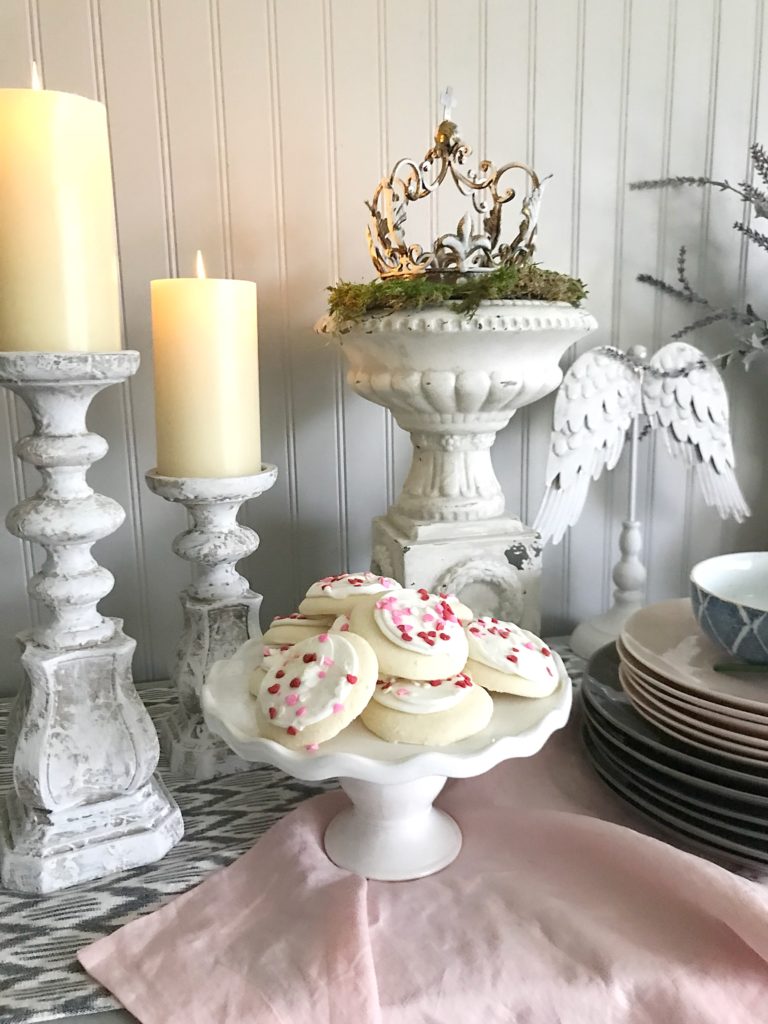 white serving platter with lots of cookies and 2 white distressed candle holders ontop of pink linen table runner and a stack of pink and grey plates next to cookies