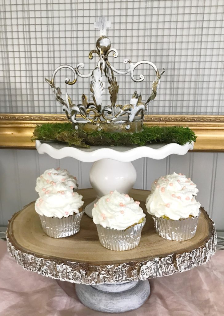 french crown on white cake stand ontop of wood cake stand with 5 white frosted cupcakes