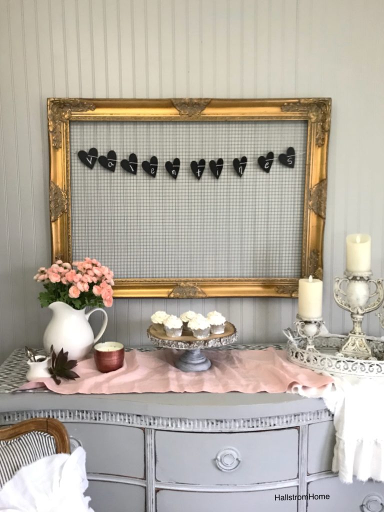 gold ornate frame with chicken wire and valentines heart banner with cupcakes and pink flowers displayed. pink linen table runner and 3 candle holders