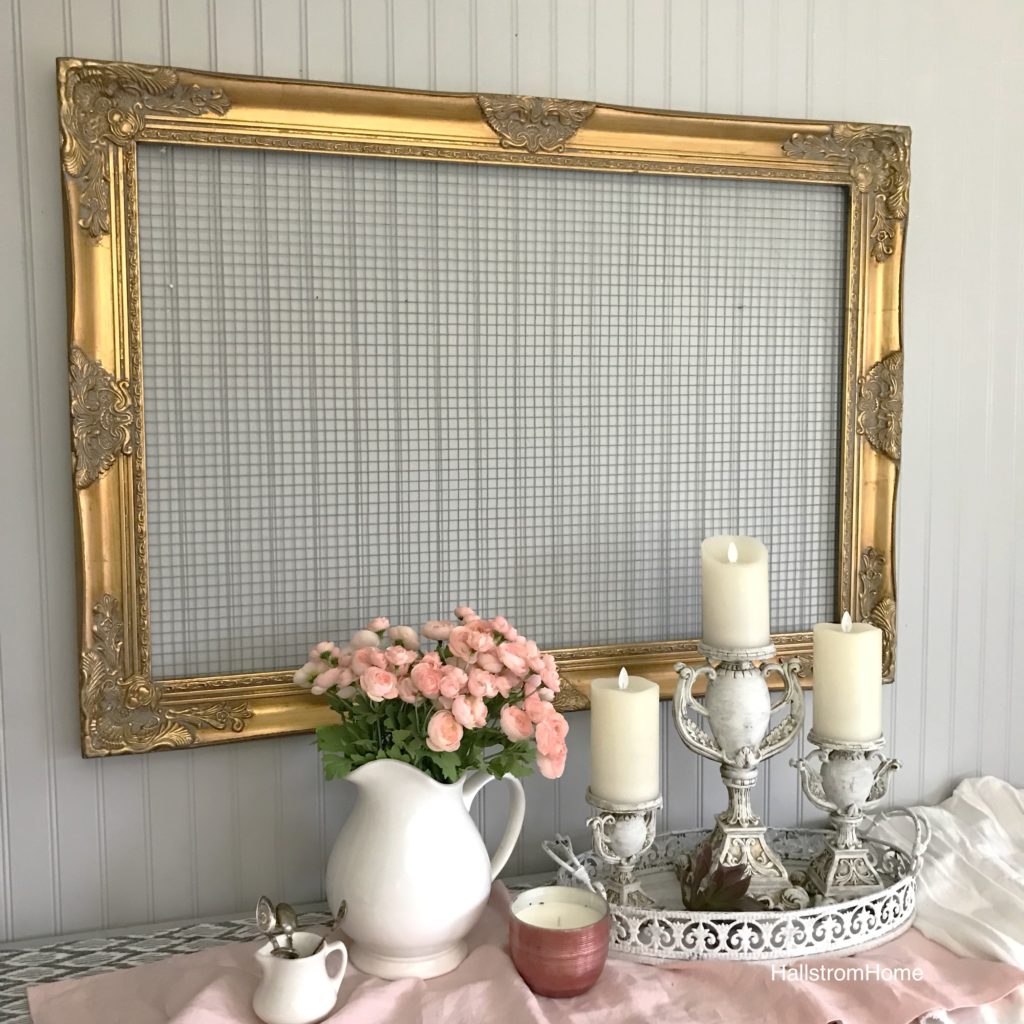 Gold ornate frame with chicken wire hanging. white pot of pink flowers and 3 candles with a pink linen table runner and pink candle