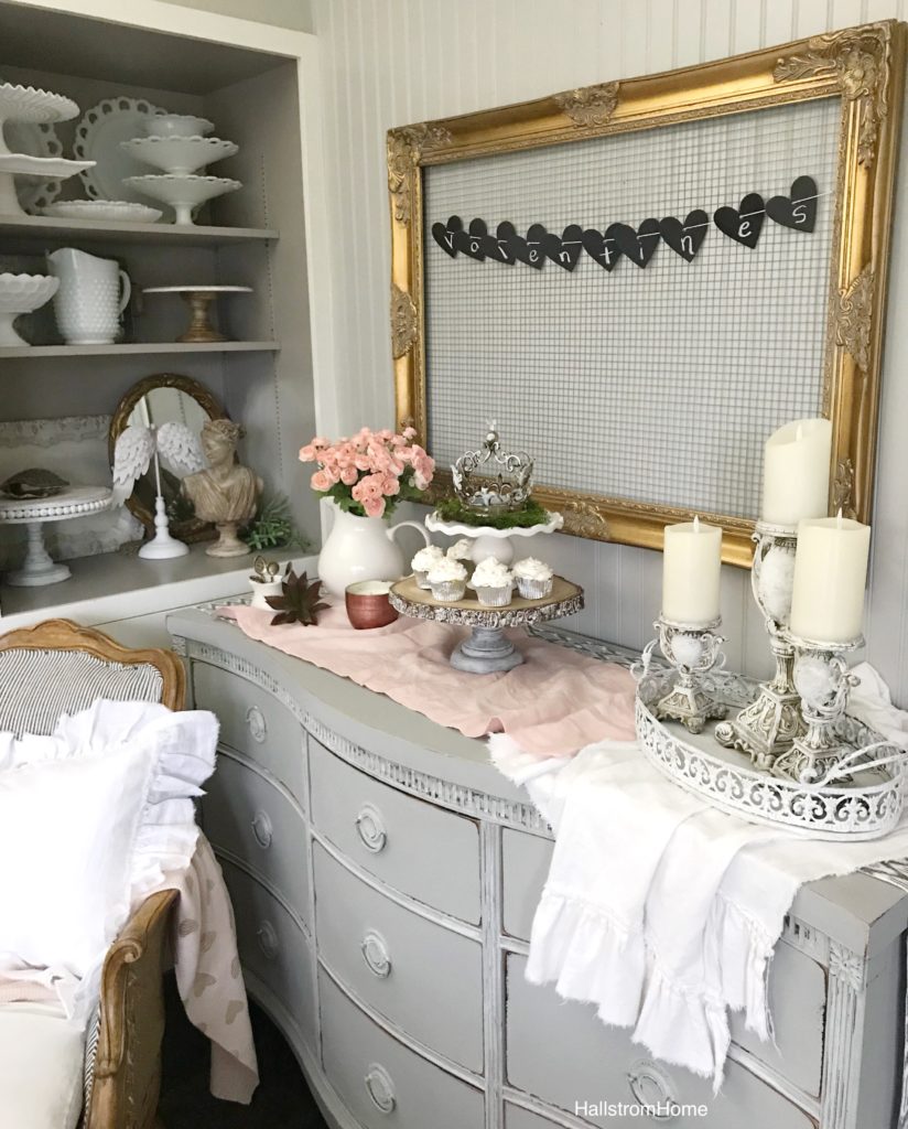 gold frame with chicken wire and valentines banner with cupcake on stand with 3 tall candle holders. 2 linen table runners and white vase with pink flowers