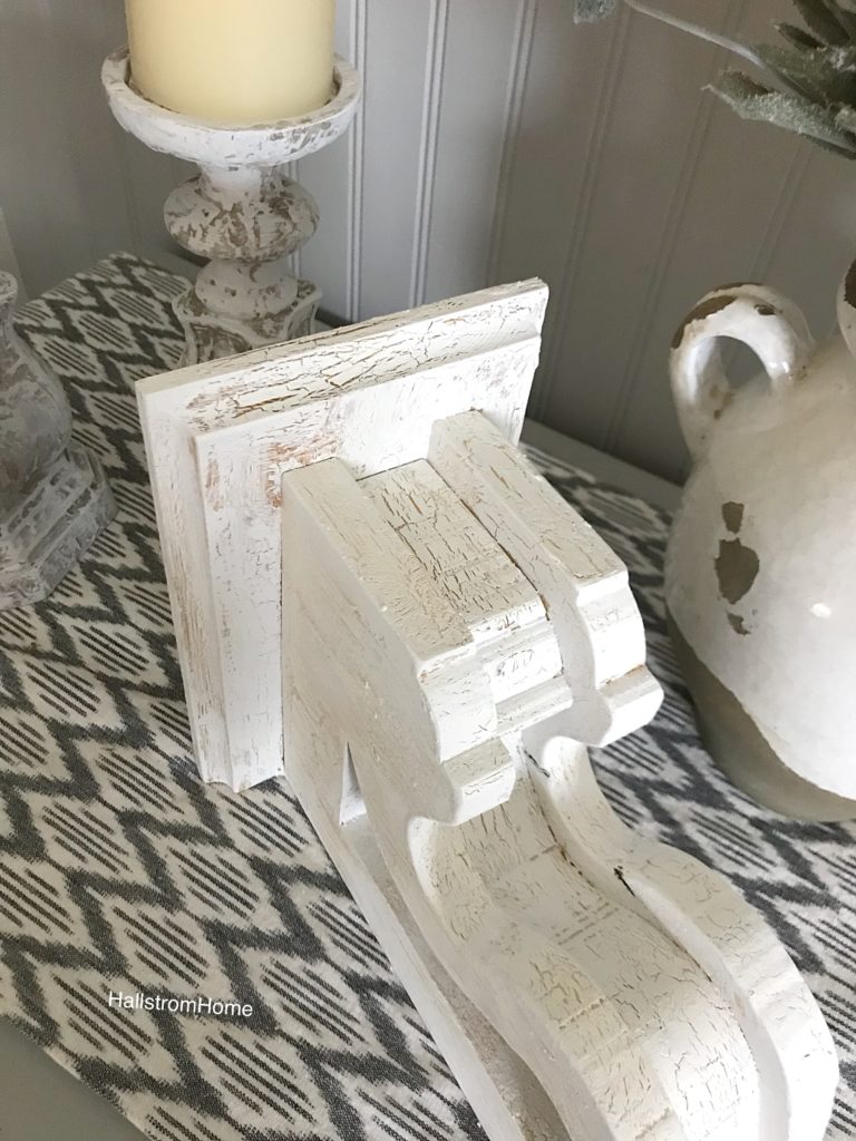 chippy painted white corbel on a patterned blue table runner and 2 white candle holders on the left and a white pot on the right