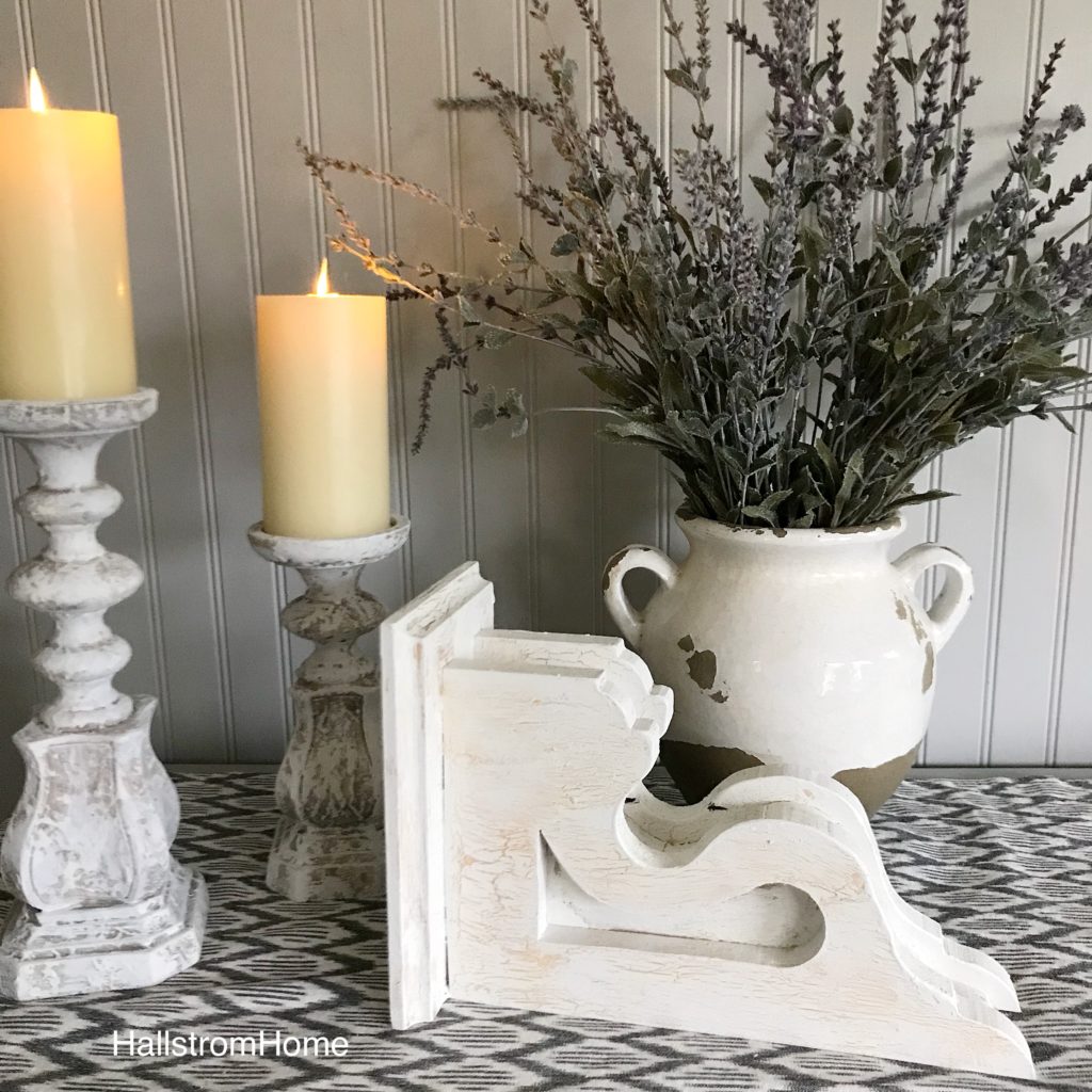 How to Chippy Paint Corbels white corbel on its side with 2 white distressed candle holders with cream candles and white pot with lavendar