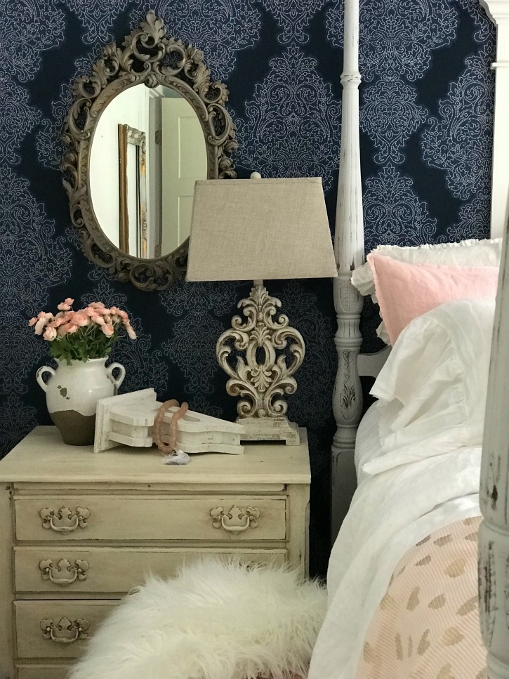 blue wall paper with brown ornate mirror hanging and night stand with ornate white table lamp and white vase with pink flowers and white chippy corbel with pink glass beads on it and white fur blanket on stool below