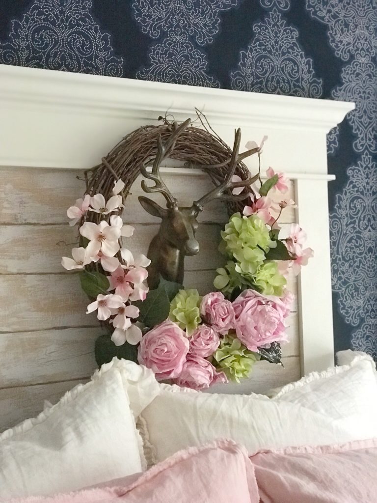 blue wallpaper and wreath hanging infront of covered window. wreath with pink and green flowers and gold deer head in the middle