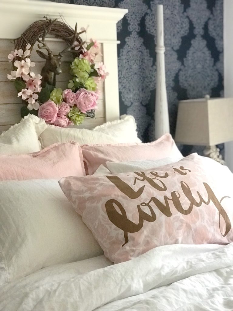bed with 4 white linen pillows and 2 pink linen pillows. accent pillow is pink and says life is lovely behind bed is wreath with pink and green flowers and gold deer head in middle