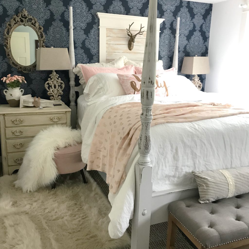 blue wall paper with ornate mirror on left of bed night stand with lamp shade and bouquet of pink flowers and a pink foot stool with white fur blanket. pink and white pillows on bed with white bedding and pink knit blanket on bed with gold hearts