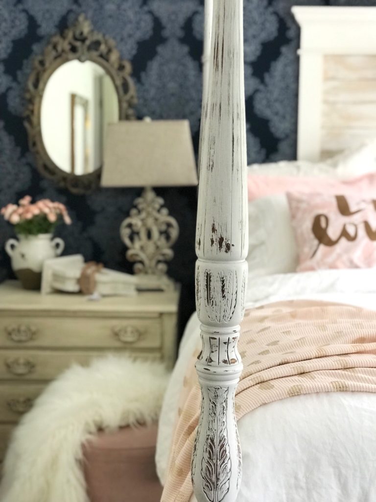 bedroom bed post in middle of picture with mirror in the background to the left with an ornate table lamp on night stand with vase of pink flowers and pink foot stool with white fur blanket. on right of bed post is bed with 4 pillows and pink throw blanket.