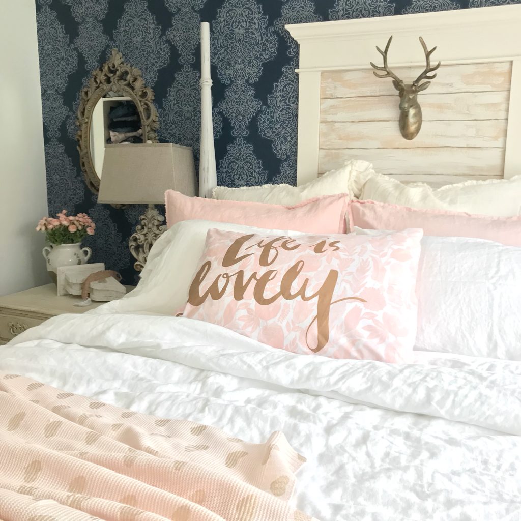 bedroom with blue wall paper and window with gold deer head behind bed. bed with 4 white linen pillows and 2 pink linen pillows and one accent pink pillow that says life is lovely 