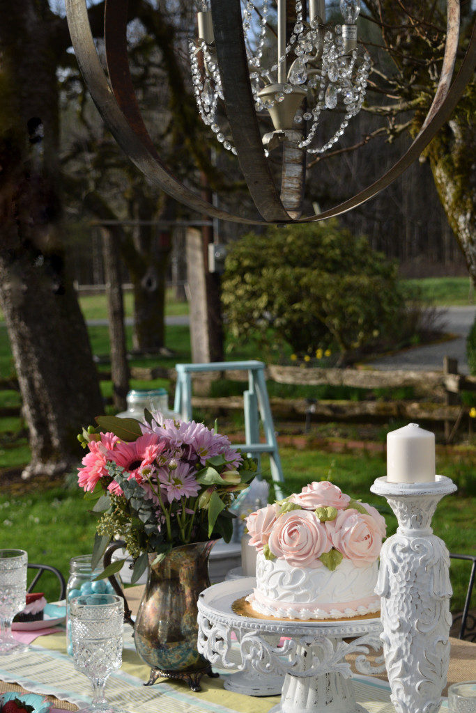 Easter Table Setting with Mini Cheesecakes white cake with pink frosted roses. tall white candle holder. outdoors with hanging chandelier silver vase on table with pink and purple flowers