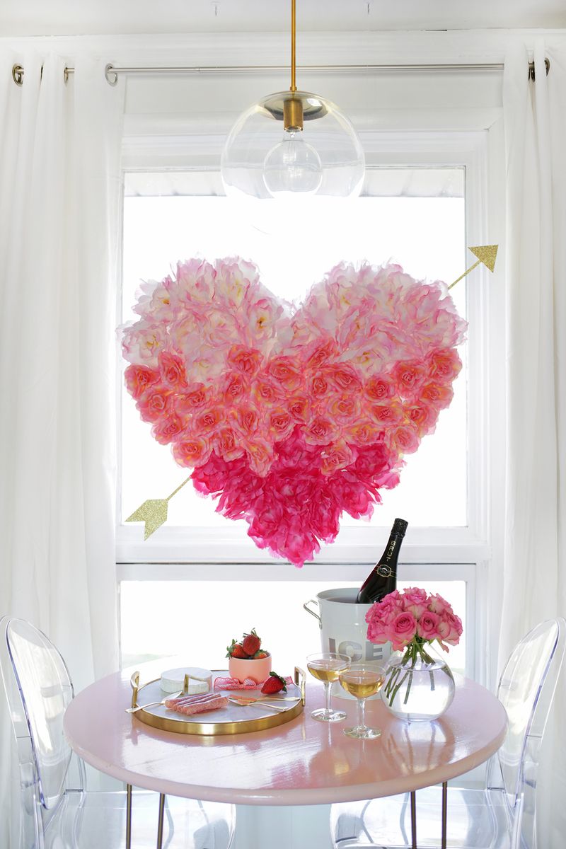 large pink flower heart with gold arrow coming out hanging above small 2 seat table. clear vase with pink flowers 2 glasses filled and an ice bucket with bottle of drink