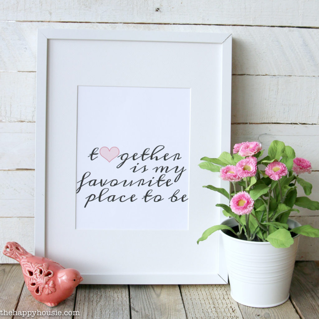white picture frame with the saying together is my favourite place to be. infront of frame is small pink ceramic bird and white pot with pink flowers