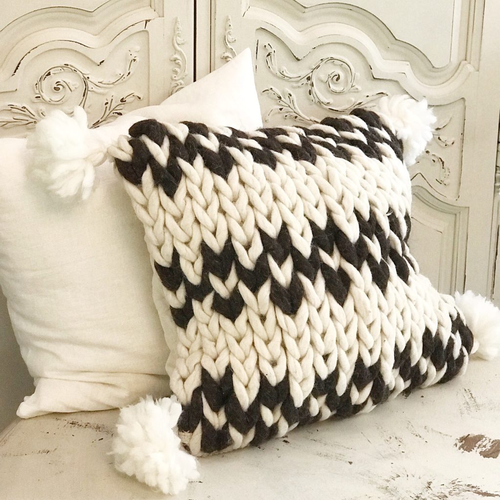 white and black chunky wool knit pillow with white tassels on corner leaning against white linen pillow