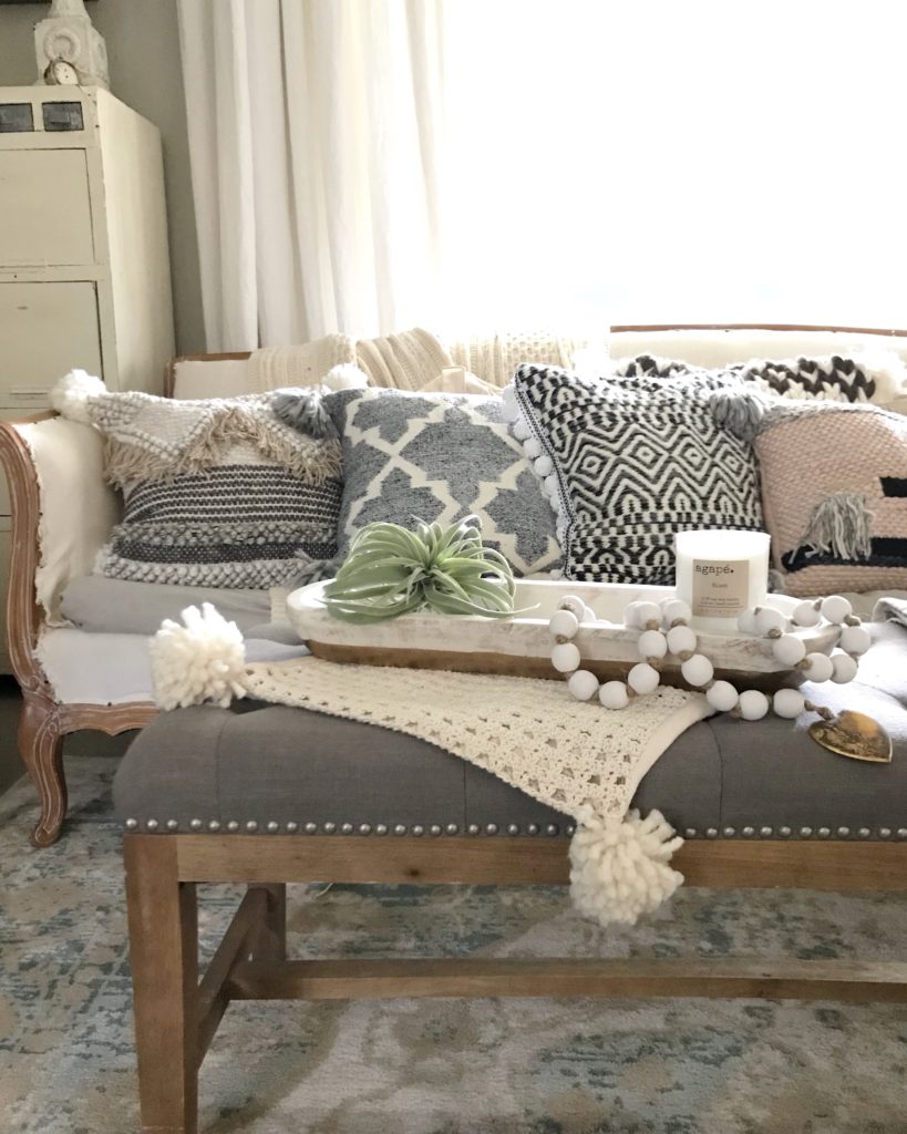 white couch with 5 chunky knit pillows and gray cushion foot rest. with gold and white dough bowl filled with wood bead garland and candle