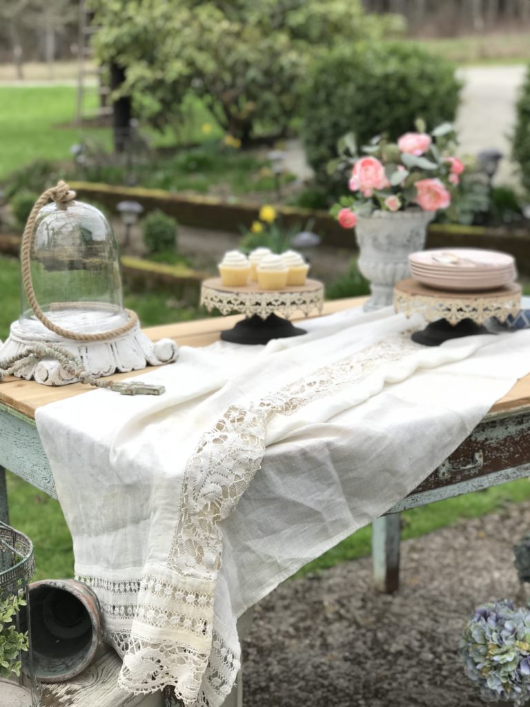 white linen sheet on farmhouse table. with 2 cake stands one with 4 white cupckaes and one with a stack of pink plates.