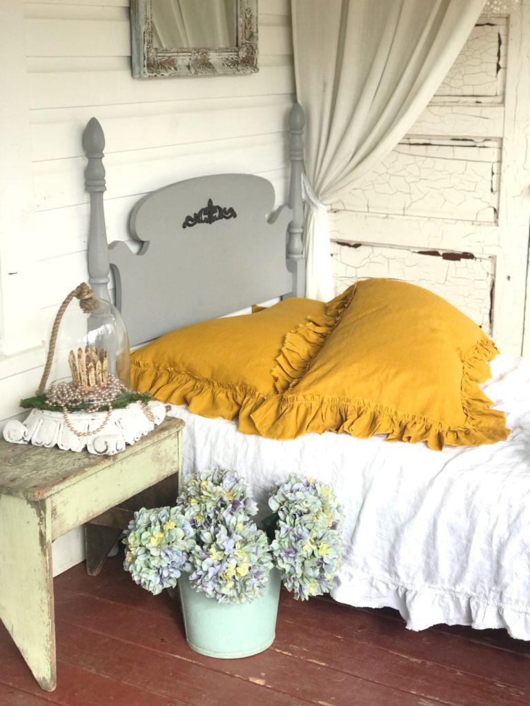 2 yellow linen pillows on white bed with gray twin bed frame. and hydrangeas on ground in blue bucket