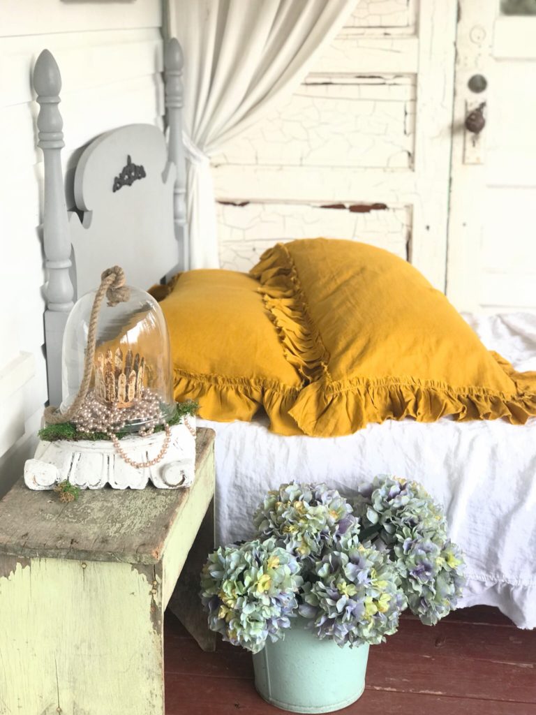 Yellow linen shams on white bed and gray twin bed frame. hydrangea in blue bucket on ground and green antique bench with white pedestal on top