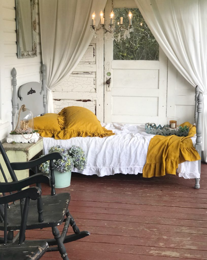 Incorporating Mustard Yellow in Shabby Chic Style napping porch with yellow pillows and bed blanket with white bedding. gray twin bed frame and chandelier over bed