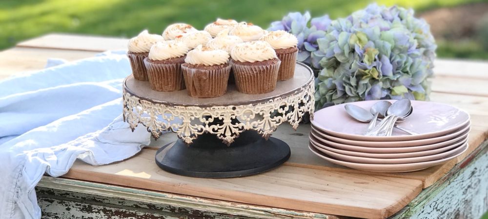 white rustic cake stand with 10 white frosted cupcakes and stack of pink plates with silver spoons ontop. bouquet of blue hydrangeas on a blue chippy farmhouse table
