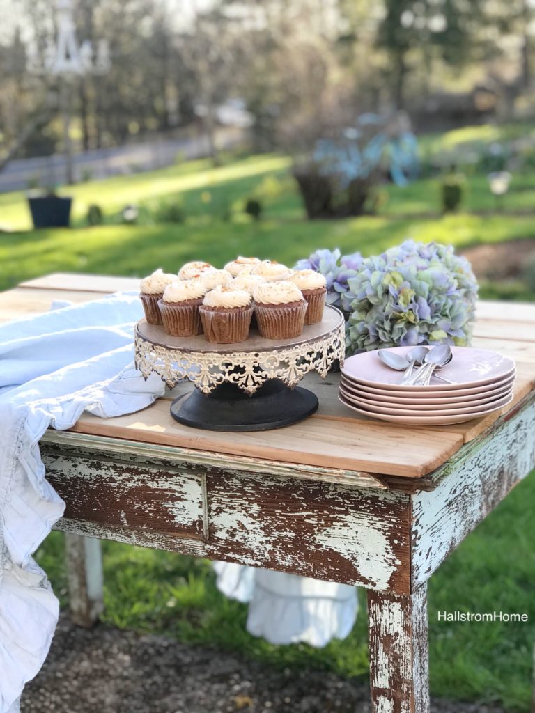 Amazing Carrot Cupcakes with Sweet Orange Frosting with white rustic cake stand with 10 white frosted cupcakes and stack of pink plates with silver spoons ontop. bouquet of blue hydrangeas on a blue chippy farmhouse table