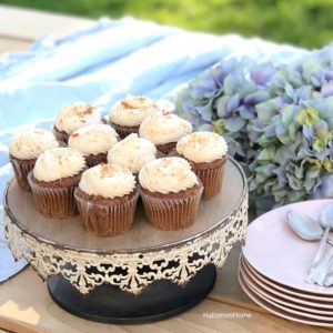 white rustic cake stand with 10 white frosted cupcakes and stack of pink plates with silver spoons ontop. bouquet of blue hydrangeas