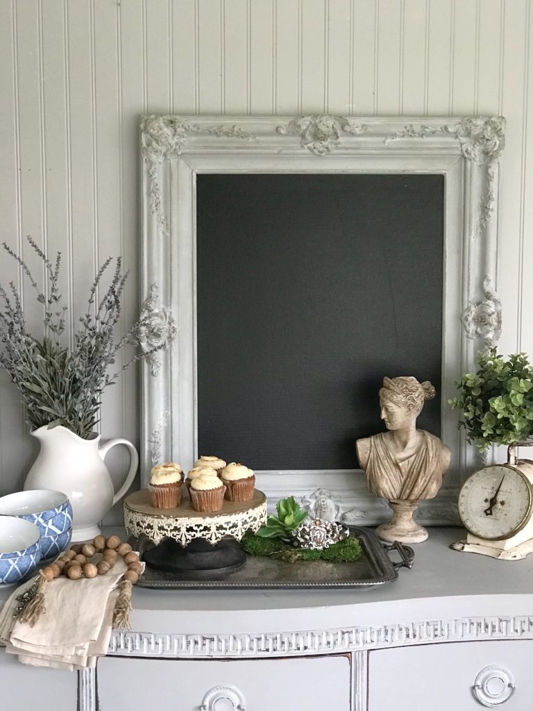 gray vintage chalkboard with white vase filled with lavender and cupcake tray with 4 white cupckaes and roman but