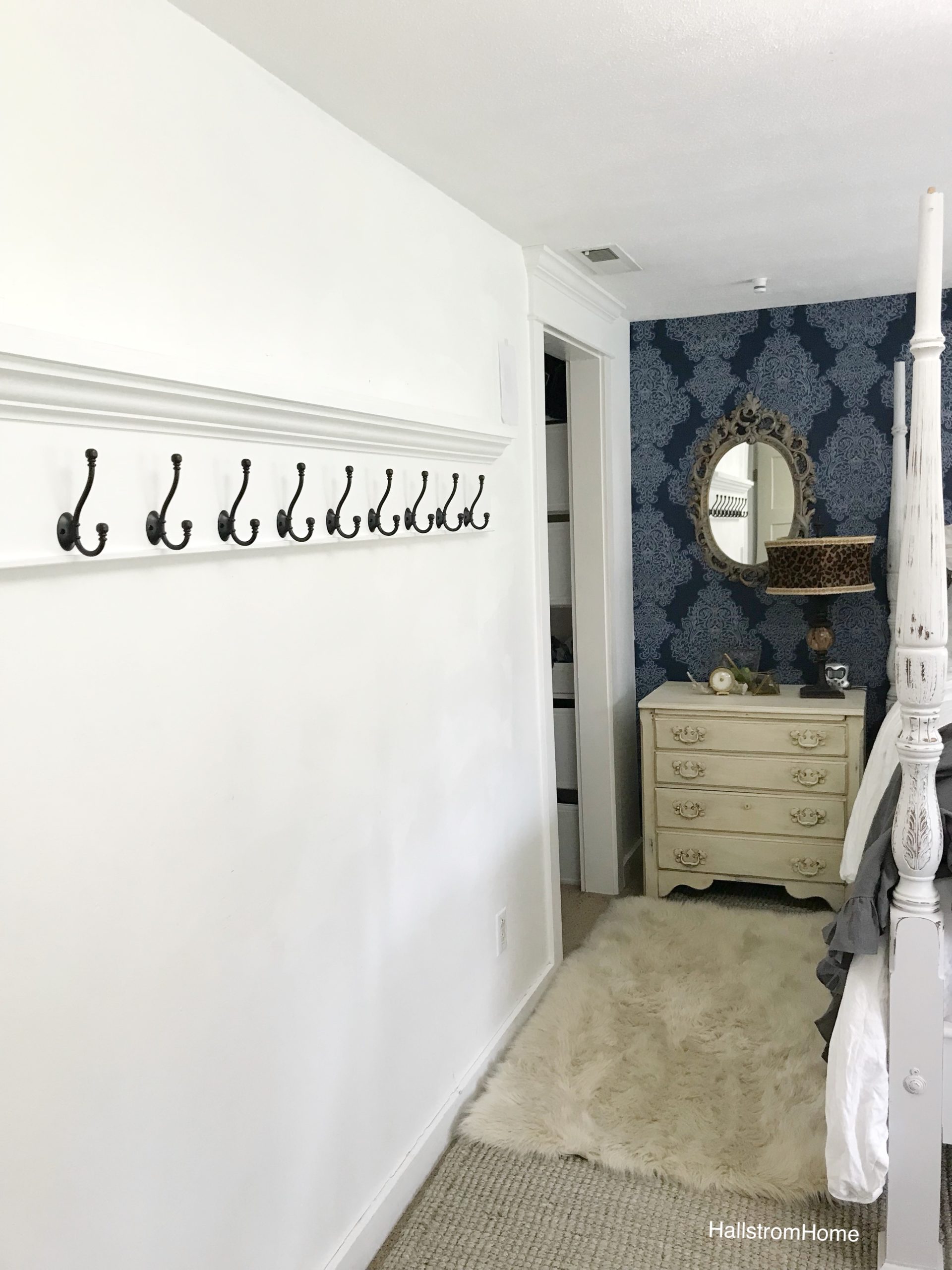 extra long coat rack hanging on white wall. other wall blue wallpaper with white dresser and oval mirror with leapord lampshade and fur rug