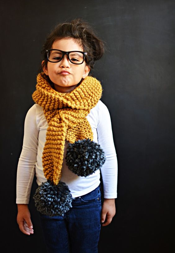 little brown haired girl with glasses and chunky yellow scarf with black pom pom on each end