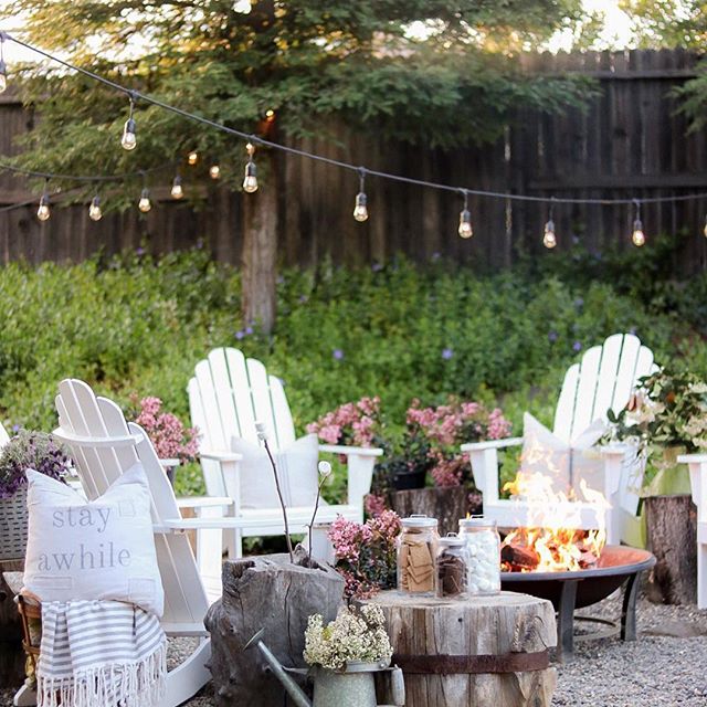 white lawn chairs around metal fire pit. wood side table with pink flowers and small jars filled with smores