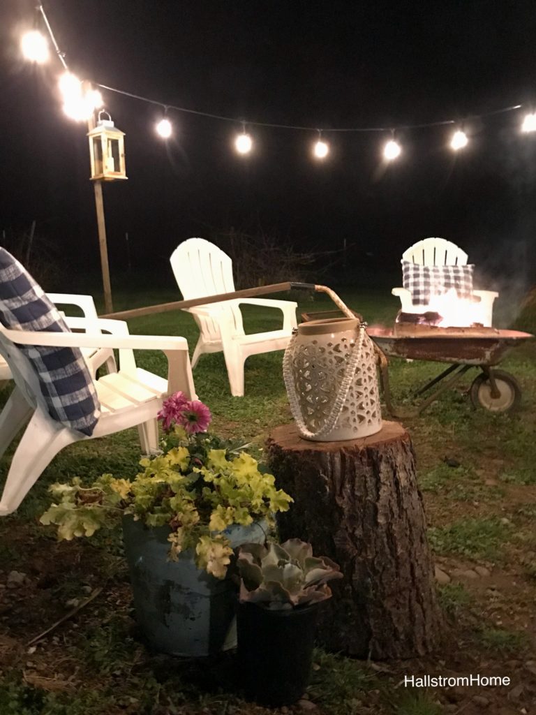 The Girl Who Built a Beautiful Fire Pit white lawn chairs around heelbarrow lit fire. lights draped around border seating area