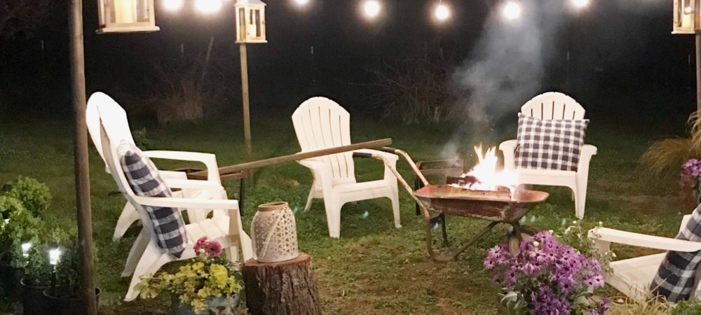 The Girl Who Built a Beautiful Fire Pit white lawn chairs around heelbarrow lit fire. lights draped around border seating area