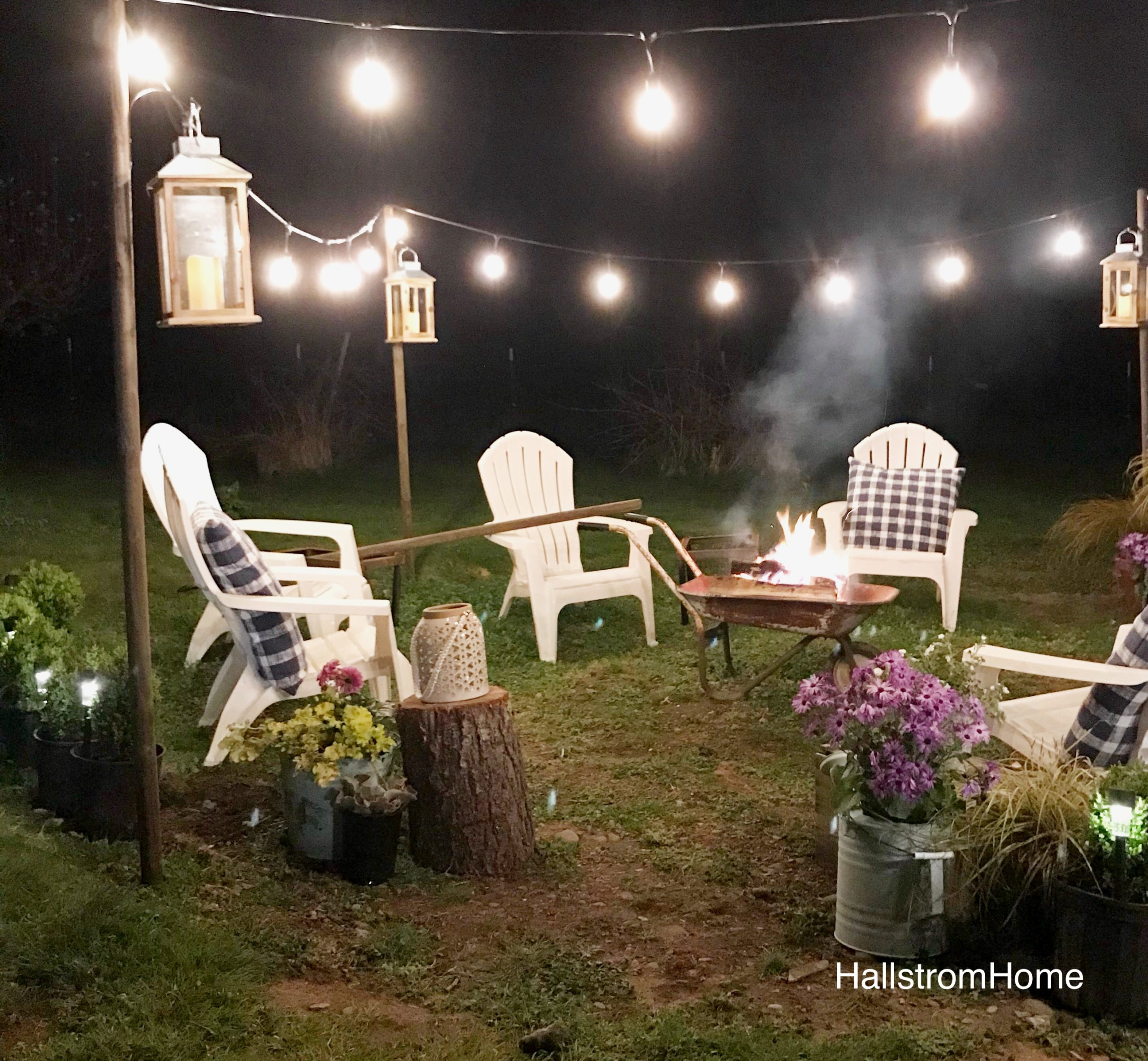 The Girl Who Built a Beautiful Fire Pit white lawn chairs around heelbarrow lit fire. lights draped around border seating area