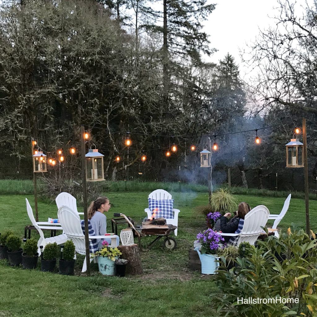 The Girl Who Built a Beautiful Fire Pit white lawn chairs around heelbarrow lit fire. lights draped around border seating area