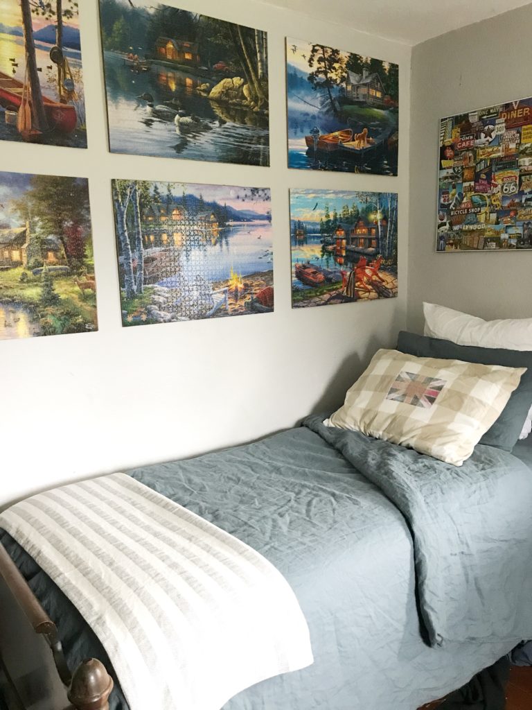blue sheets on twin bed with striped blanket at foot and 7 puzzles on the wall