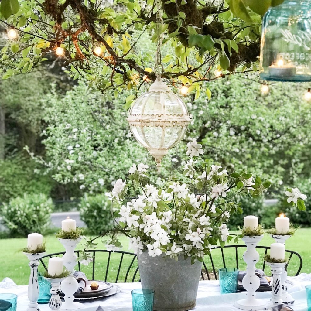 Outdoor Farmhouse Dining Made Easy white linen table cloth with 6 white candle holders with white candles and greenery. silver bucket holding white and green flowers round glass chandeiler hanging above in trees