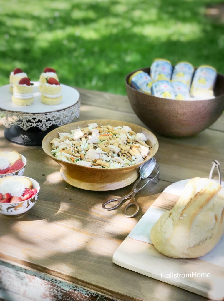 bread on cutting borad, bowl of salad, copper bowl of canned drinks, cake stand of 4 cupckaes on wooden table