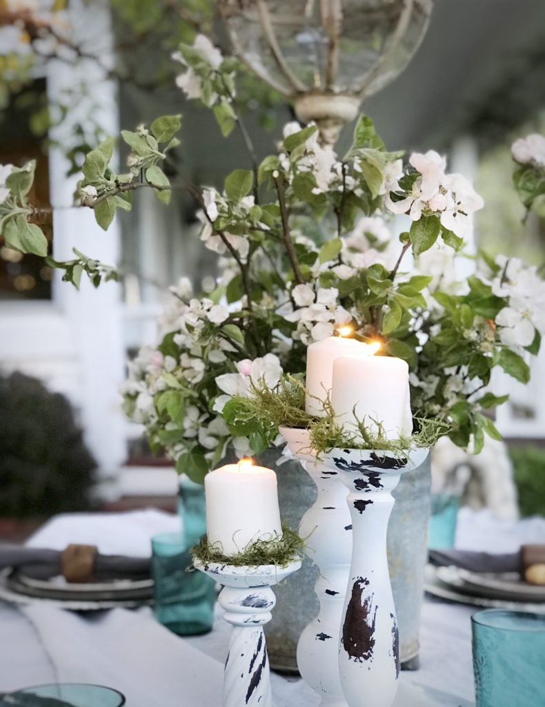 3 white distressed candle holder on outdoor table. silver bucket with white flowers and greenery