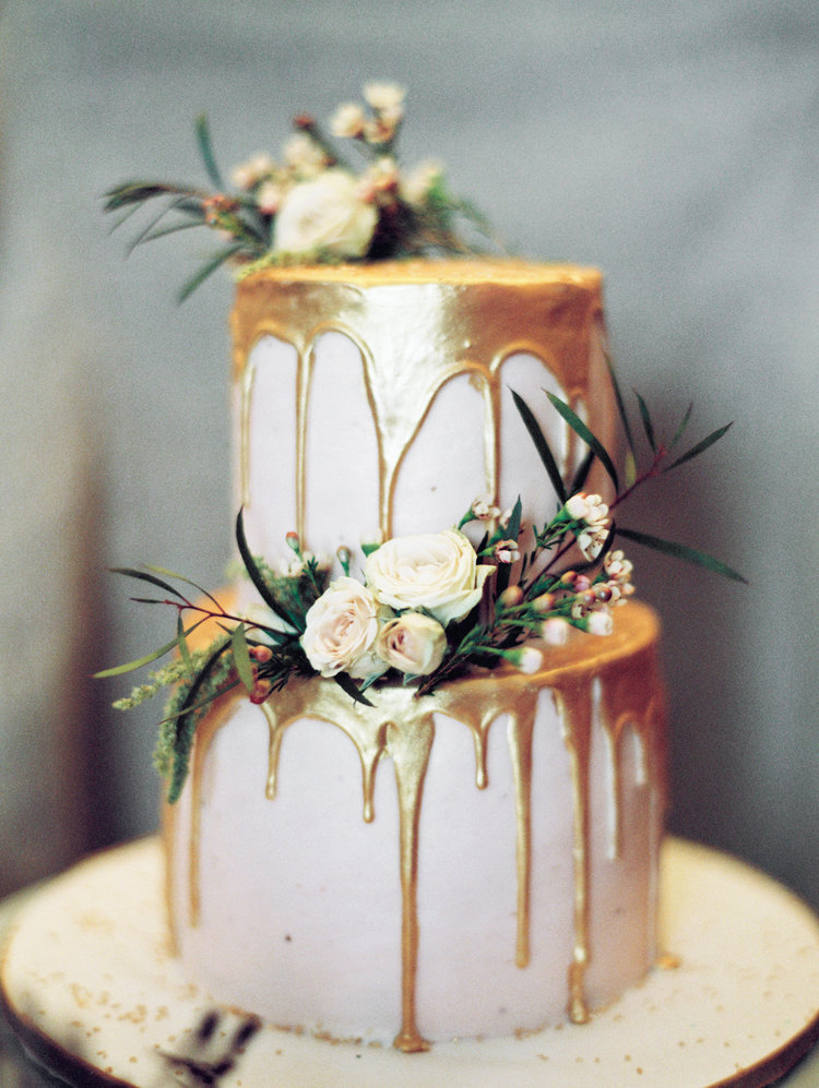 2 tier white cake with gold frosting dripping down it and white roses and greenery on it