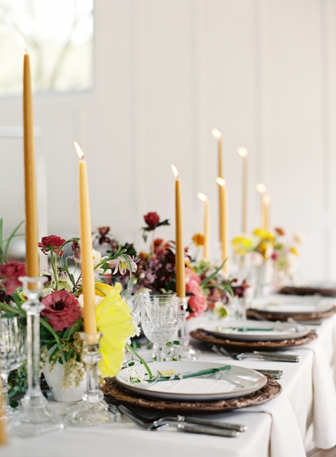 tall yellow candles with pink and green flowers in the centerpiece of white table with bronze chragers