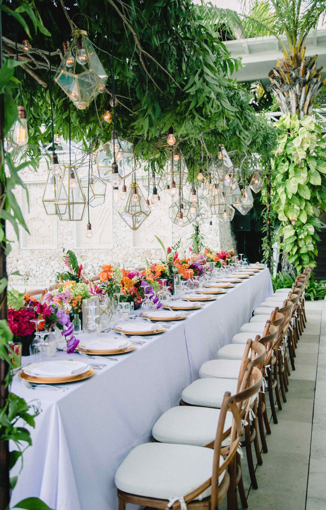 long white table with gold chargers and bright orange red purple flower centerpieces with gold light hanging above