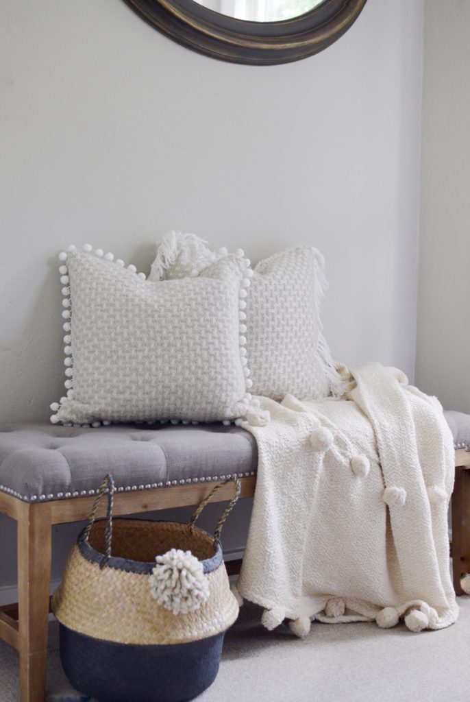 grey tufted bench with 2 pom pom pilows and a cream pom pom blanket with basket on the ground