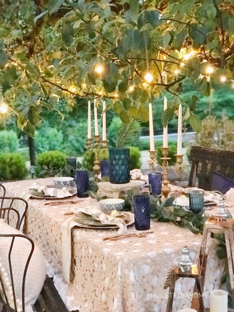 lace table cloth outside with blue cups and vase with gold candelabras and lights above on tree