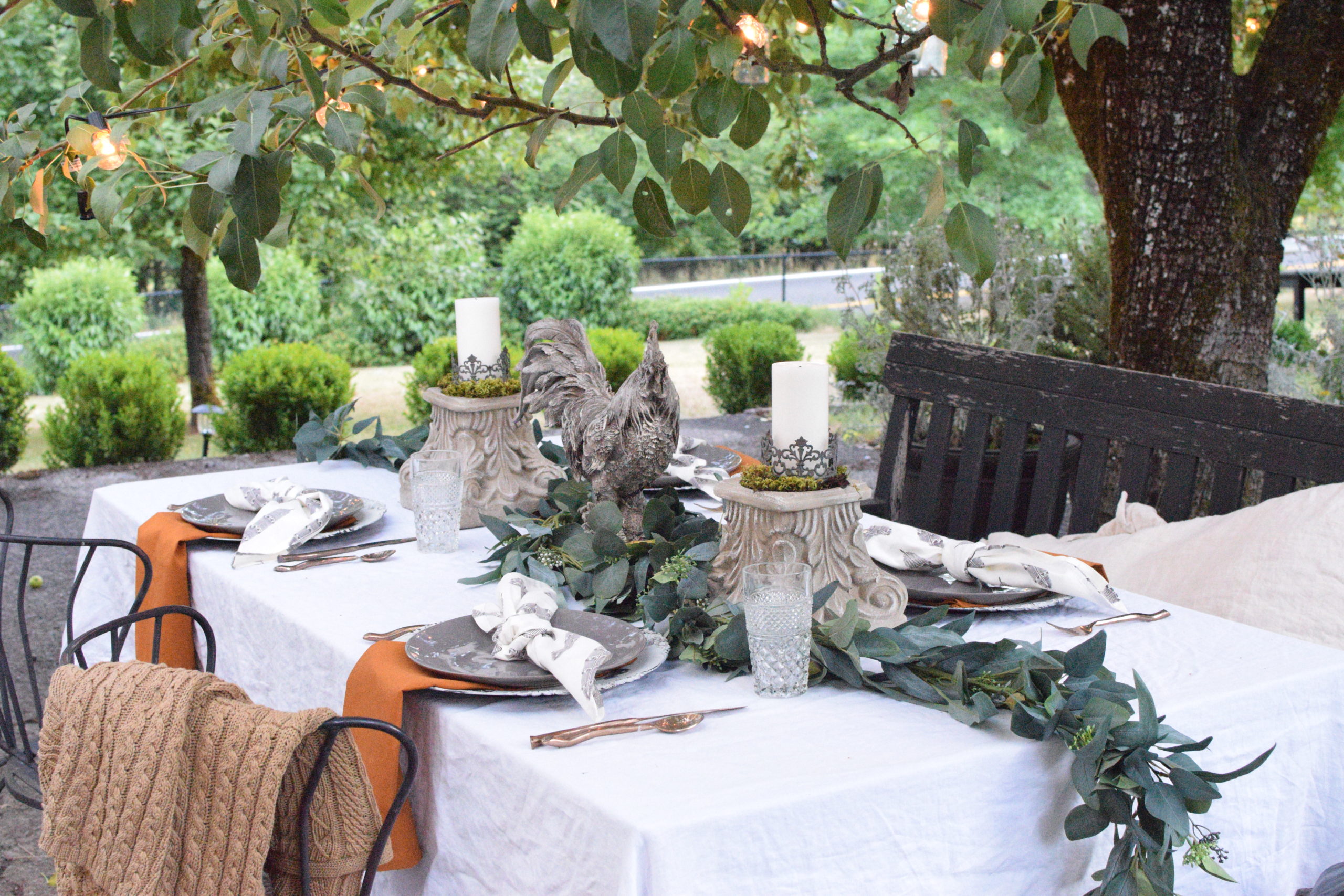 white linen table cloth qith greenery in middle and 2 candle pedestals with rooster in centerpiece