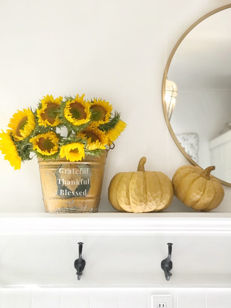 mantel with 2 yellow pumpkins and yellow metal bucket with sunflowers and gold round mirror hanging