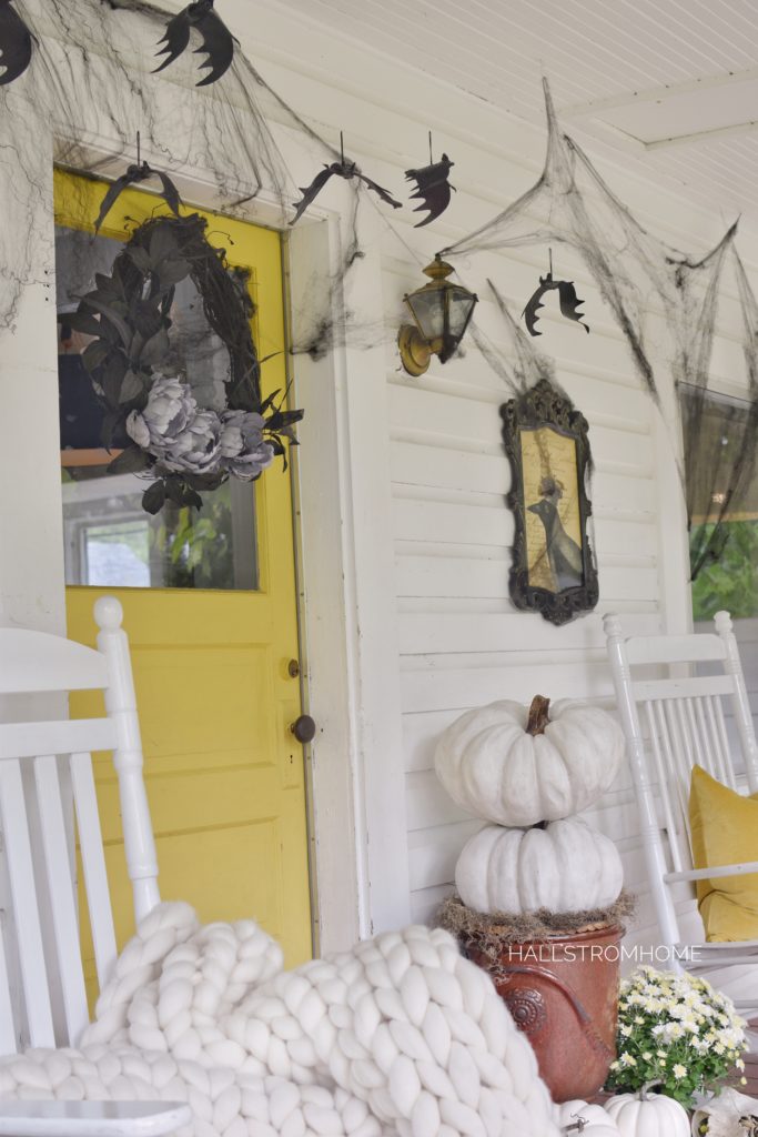 yellow door with black wreath and black webs around door with bat hanging and a stack of white pumpkins