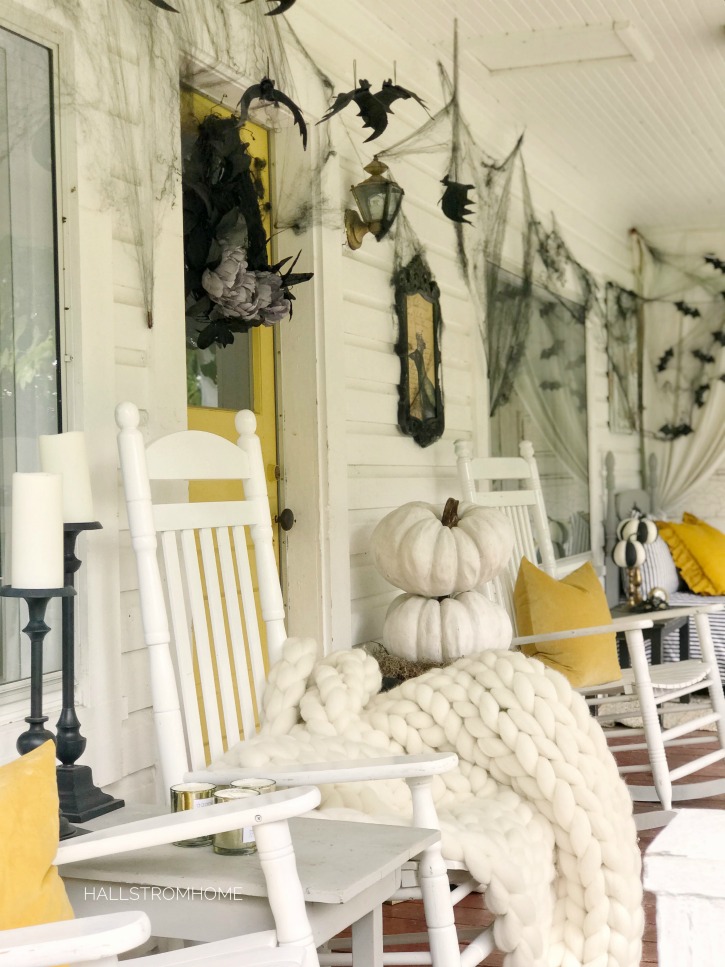 yellow door with 3 white rocking chairs on porch with hanging bats and cobwebs wiht pumpkins and whte chunky blanket on one rocking chair